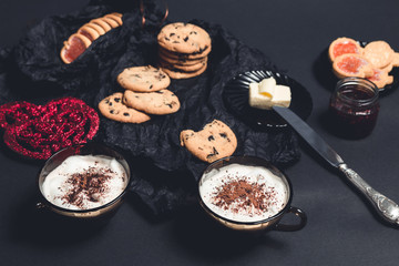 Romantic breakfast. Two Cups of coffee, cappuccino with chocolate cookies and biscuits  near red hearts on black table background. Valentine day. Love. Top view.