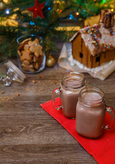 Two glasses of cocoa on wooden table and Gingerbread house, cookie jar and christmas tree with toys and garland on background.