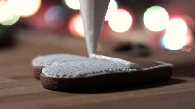 Female hand decorate Gingerbread white heart cookie with food icing on the table, close up. Candle, pine cone, mandarin, canella, bokeh in backgound.