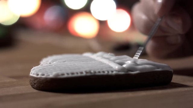 Female hand decorate Gingerbread white heart cookie with food icing on the table, close up. Candle, pine cone, mandarin, canella, bokeh in backgound.