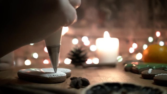 Female hand decorate Gingerbread white heart cookie with food icing on the table, close up. Candle, pine cone, mandarin, canella, bokeh in backgound.
