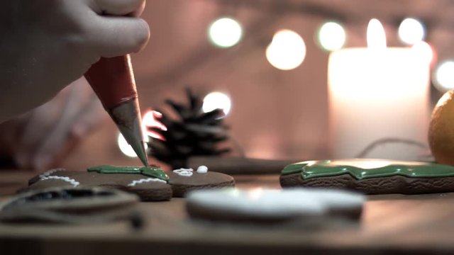 Female hand decorate Gingerbread man cookie with food icing on the table, close up. Candle, pine cone, mandarin, canella, bokeh in backgound.