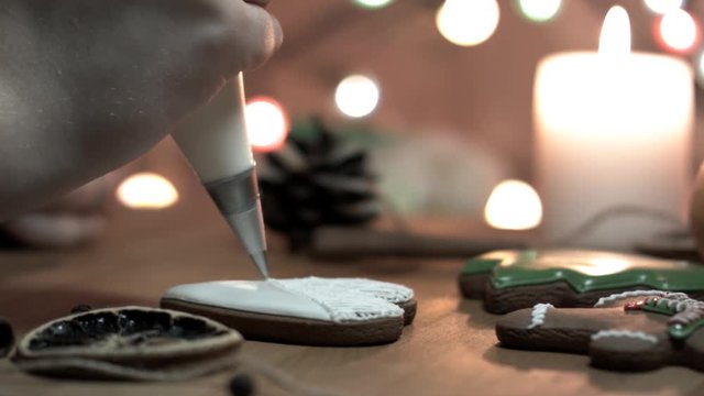 Female hand decorate Gingerbread white heart cookie with food icing on the table, close up. Candle, pine cone, mandarin, canella, bokeh in backgound.