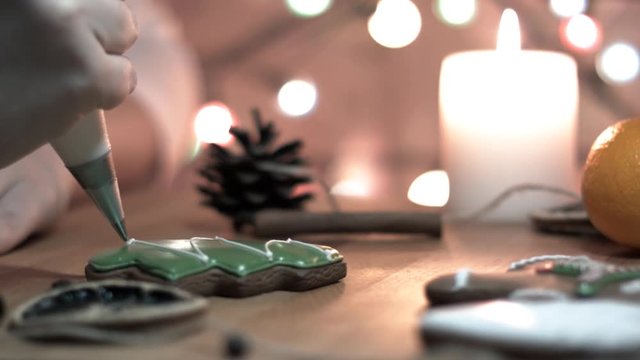 Female hand decorate Gingerbread tree cookie with food icing on the table, close up. Candle, pine cone, mandarin, canella, bokeh in backgound.