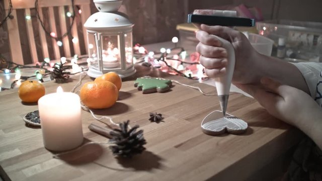 Female hand decorate Gingerbread white heart cookie with food icing on the table, close up. Candle, pine cone, mandarin, canella, bokeh in backgound.