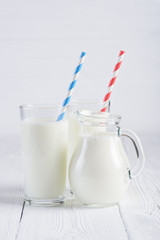 Glass of milk with stripped blue and red paper straws and jug of milk on white wooden table still life