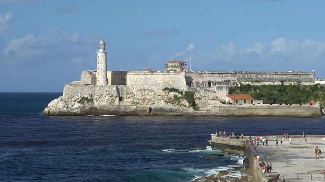 View on big stone lighthouse into sea bay which is placed in fortress at downtown of Havana. People are walking by seafront.