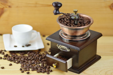 Old-style manual coffee grinder on a rustic table with wooden planks. Around the mill are scattered coffee beans. Near the mill stands empty white cup.
