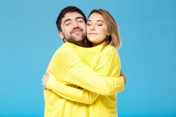 Young beautiful couple in one yellow sweater embracing over blue background.