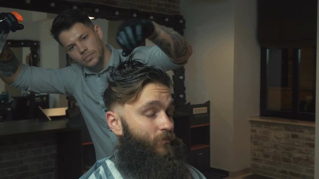 Barber with dark hair and tattoo wearing white shirt and black gloves doing a haircut for brutal client with scissors at barber shop