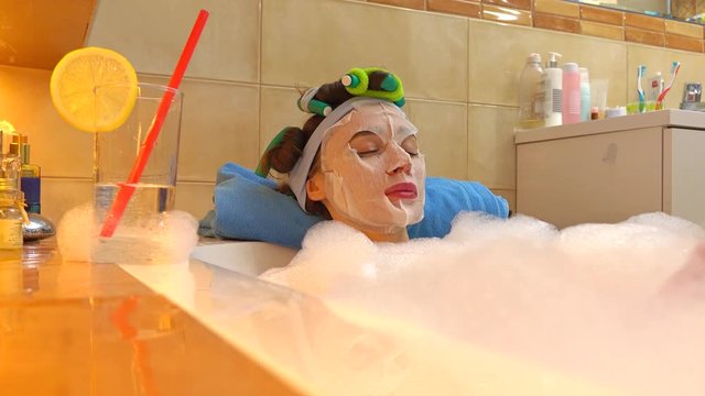 Beautiful brunette woman wearing cosmetic face mask relaxes in foamy bath. Beauty treatment at home. 4K shot