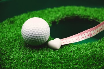 golf ball and white heart shape on green grass at hole with measuring tape.