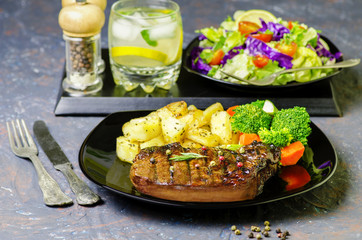 Delicious juicy beefsteak with fried potatoes and steamed vegetables served in black plate with fresh green salad and drink..