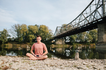 man practices yoga on the river bank near the old bridge
