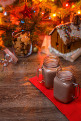 Two glasses of cocoa on wooden table and Gingerbread house, cookie jar and christmas tree with toys and garland on background.