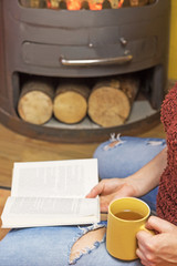 Woman is sitting down and holding yellow tea mug and open book. Burning wood stoves are in the background. Vertically. The letters in the book are intentionally blurred.