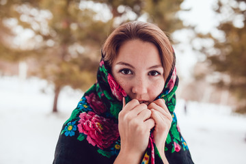 Russian beauty woman with red hair and freckles in the national scarf stands on a background of trees.