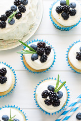 Homemade cupcakes and cake with frosting, blueberries, blackberries and rosemary leaves on white wooden background