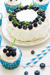 Homemade cupcakes and cake with frosting, blueberries, blackberries and rosemary leaves on white wooden background