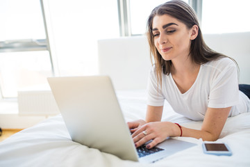 Portrait of beautiful happy young woman lying in bed and using laptop at home