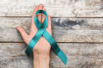 Female hand holding sea-green ribbon on wooden background. Ovarian cancer and gynecological disorders concept