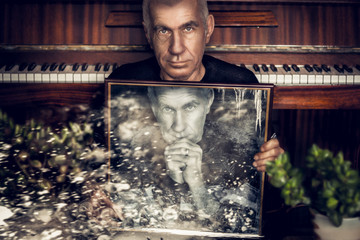 Dramatic portrait of an old grandfather with a pensive look on the background of the piano that holds the frame with his twin. He looks into the camera. Close-up portrait. Double exposure.