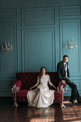 cute wedding couple in the interior of a classic studio posing at the sofa . hey kiss and hug each other, holding hands looking at each other