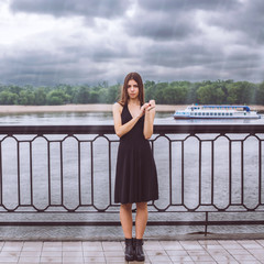 Model - cute brunette with green eyes, wearing a dress. A girl stands at the railing in the rain on a background of the river / trees. The sky is covered with clouds / storm / thundercloud.