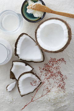 Pieces of a whole coconut cracked open on a white background. Coconut milk, water, flakes and oil are placed around the raw coconut in the center.