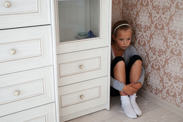 Sad little girl sitting in the corner of a room behind the cupboard