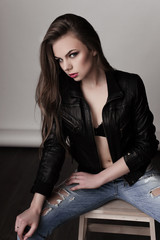 Young girl with long beautiful hair and smoky eyes wearing black leather jacket and jeans sitting on chair. Studio shot.