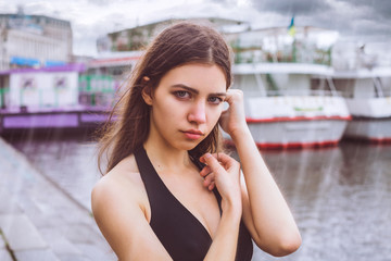 Model - cute brunette with green eyes, wearing a dress. Girl standing in the rain on a background of the river / buildings / yachts. The sky is covered with clouds / storm / storm cloud.