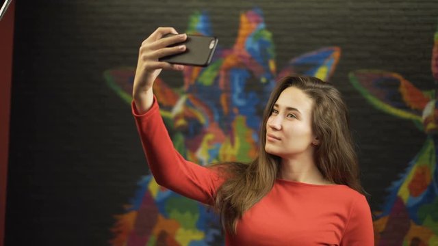 Young beautiful girl in a red dress makes selfie on your smartphone. In room.