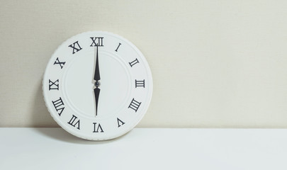 Closeup white clock for decorate in 6 o'clock on white wood desk and cream wallpaper textured background with copy space
