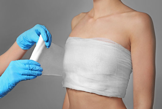 Doctor wrapping bandage around young woman's chest on grey background, closeup