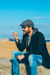 Young man sitting on coastline having conversation with voice message recorder on smartphone.
