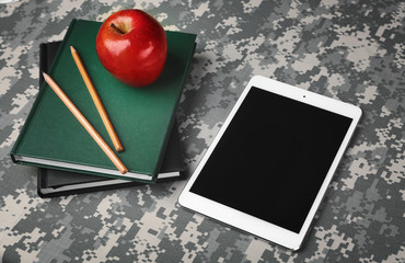 Military education concept. Tablet, books, pencils and apple on camouflage background