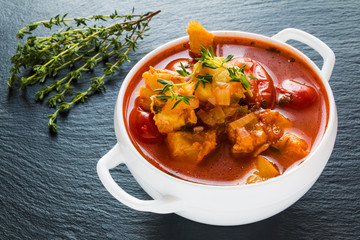 Fish soup with cod, tomato, onion, garlic and thyme in white bowl on black stone background.
