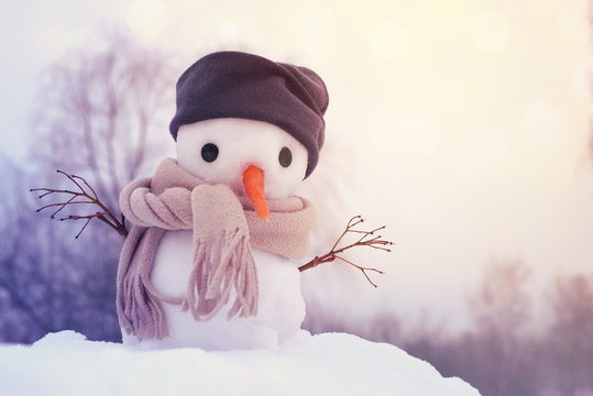Small snowman in a cap and a scarf on snow in the winter against the background of trees. Festive background with a lovely snowman. Christmas Card, copy space