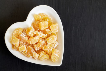 Candied ginger in heart shaped bowl on black wooden background. Top view with copy space