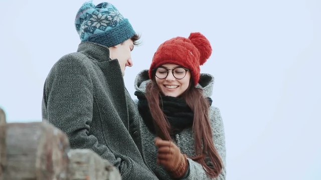 Woman with a young man in a coat and knit cap, sitting, against the gray sky, they are talking laughing.