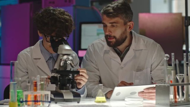  Lockdown of cheerful curly female scientist looking into microscope, showing something on tablet to bearded male colleague and discussing work 
