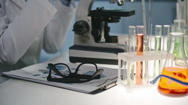 Tilt up with slow motion of young female scientist taking off her glasses and looking at sample in microscope 