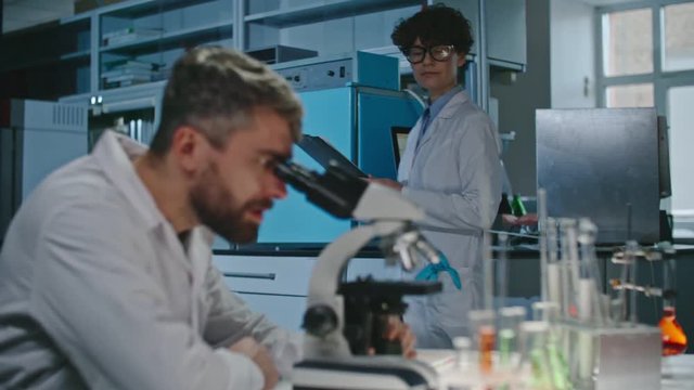 Male scientist with beard looking into microscope, then calling up his female colleague and taking from her clipboard for making notes