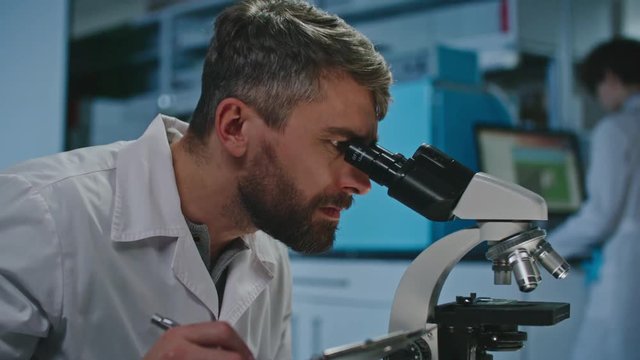 Male scientist with beard holding clipboard and looking into microscope, then calling up his female colleague and showing her