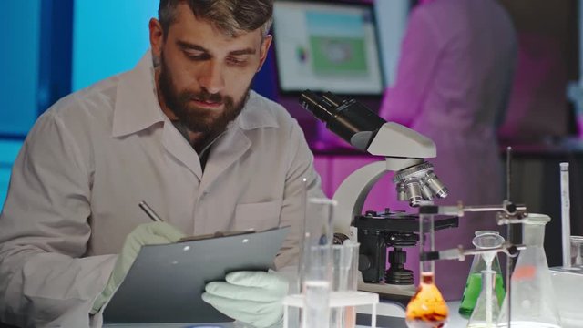 Male scientist with beard looking into microscope and making notes, then looking up and posing for camera