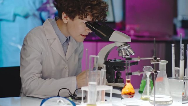 Curly female scientist in glasses looking into microscope in laboratory, then looking up and posing for camera