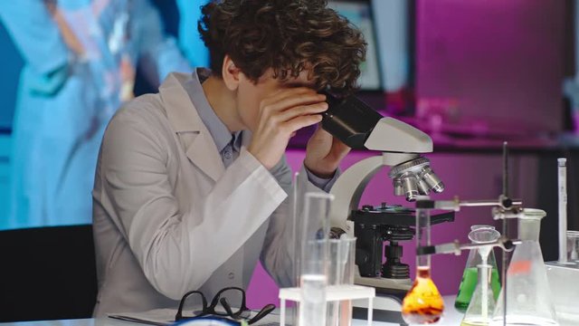 Curly female scientist taking off her glasses and looking into microscope in laboratory 