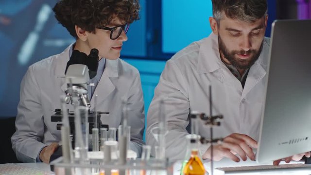 Curly female scientist in glasses looking into microscope as her male colleague typing notes on computer, and then discussing it together  