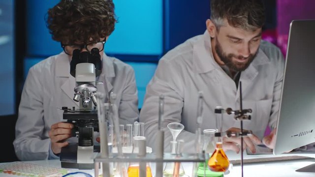 Female scientist in glasses looking into microscope and then discussing work with male bearded colleague typing notes on computer 